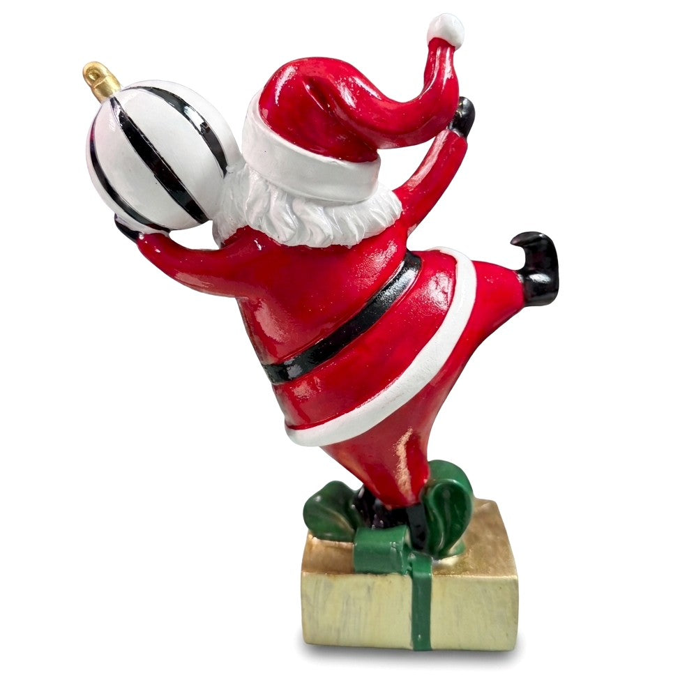 Babbo Natale in poliresina visto da dietro, in equilibrio su una gamba sul pacco regalo dorato, con palla a righe e cappello rosso